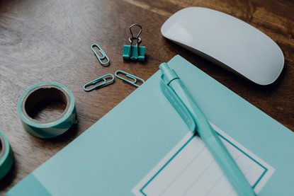 Auf dem Tisch liegen ein türkises Heft und ein türkiser Stift, türkisfarbenes Washitape, türkisfarbene Büroklammern und eine Maus der Marke Apple