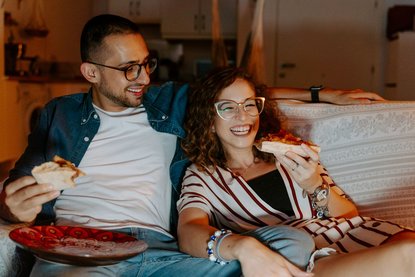 Mann und Frau sitzen auf dem Sofa, essen Pizza und schauen etwas.